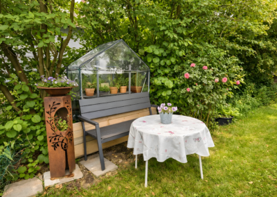 Gemütlicher Garten-Sitzplatz mit Mini-Gewächshaus, Blumen, Rosen und dekorativem Metallständer bei der Ferienwohnung Jaeger in Viereth bei Bamberg.
