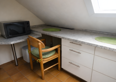 Kleiner Küchenbereich unter der Dachschräge mit Mikrowelle, Arbeitsplatte, Stuhl und Dachfenster in der Ferienwohnung Jaeger in Viereth bei Bamberg.